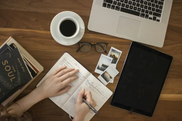 Title: Writer drafting notes in a notebook beside a laptop and a cup of coffee - Description: Writer drafting notes in a notebook beside a laptop and a cup of coffee
