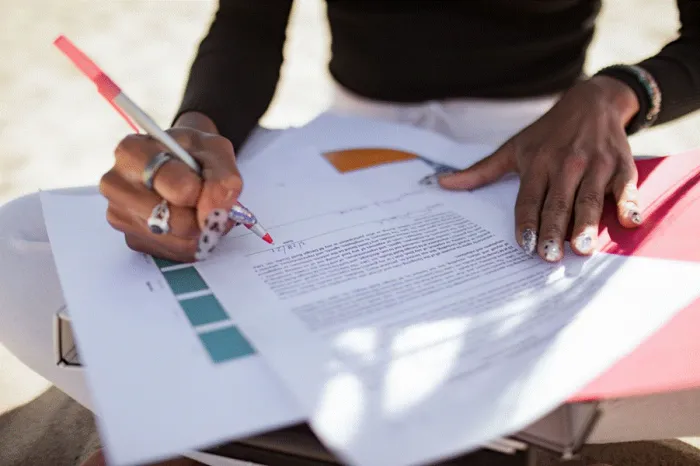 Title: Hands marking up a printed article draft with a red pen at an outdoor table - Description: Hands marking up a printed article draft with a red pen at an outdoor table