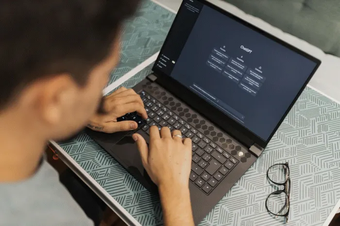 Title: Person wearing glasses typing on a laptop with the ChatGPT interface open - Description: Person wearing glasses typing on a laptop with the ChatGPT interface open