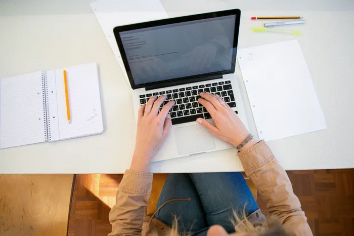 Title: Overhead view of a person typing on a laptop beside an open notebook and pens - Description: Overhead view of a person typing on a laptop beside an open notebook and pens