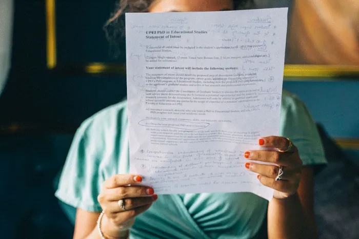 Title: Woman reading a printed page covered in handwritten corrections, with a laptop on her knees - Description: Woman reading a printed page covered in handwritten corrections, with a laptop on her knees